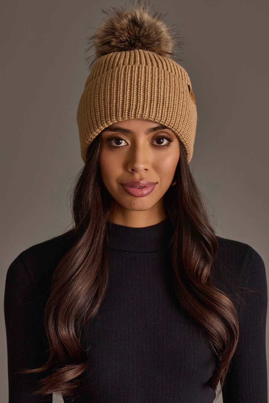 A woman with long, wavy brown hair wears the Panache Apparel Collection Tan Knit Pom Hat and a black ribbed sweater, looking directly at the camera against a plain gray background.