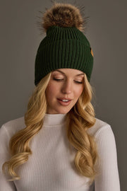 A woman with long blonde hair is wearing the Panache Apparel Collection Dark Green Knit Pom Hat, featuring a faux fur pom-pom, paired with a white ribbed turtleneck as she looks down against a plain gray background.