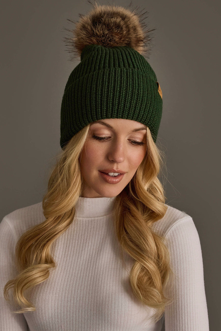A woman with long blonde hair is wearing the Panache Apparel Collection Dark Green Knit Pom Hat, featuring a faux fur pom-pom, paired with a white ribbed turtleneck as she looks down against a plain gray background.