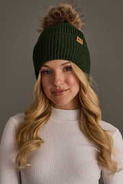 A woman with long blonde hair wearing the Panache Apparel Collection Dark Green Knit Pom Hat and a white ribbed turtleneck sweater stands against a plain gray background.