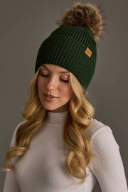 A woman with long blonde hair wears the Panache Apparel Collection’s Dark Green Knit Pom Hat and a light ribbed turtleneck, looking down with a soft smile against a neutral background.
