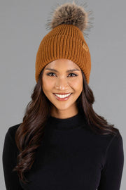 A woman with long brown hair wears the Panache Apparel Collection Camel Knit Pom Hat, featuring a stylish fur pompom, along with a black turtleneck sweater. She is smiling while standing against a plain gray background.