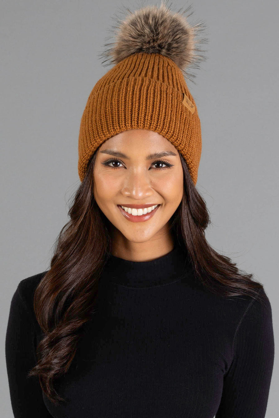 A woman with long brown hair wears the Panache Apparel Collection Camel Knit Pom Hat, featuring a stylish fur pompom, along with a black turtleneck sweater. She is smiling while standing against a plain gray background.