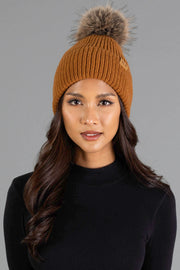 A woman with long dark hair wears the Panache Apparel Collection’s Camel Knit Pom Hat, featuring a fur pom-pom, paired with a black top, standing against a plain gray background.