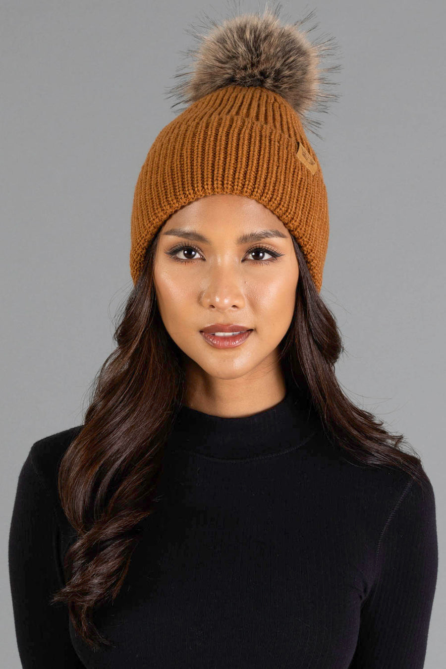A woman with long dark hair wears the Panache Apparel Collection’s Camel Knit Pom Hat, featuring a fur pom-pom, paired with a black top, standing against a plain gray background.