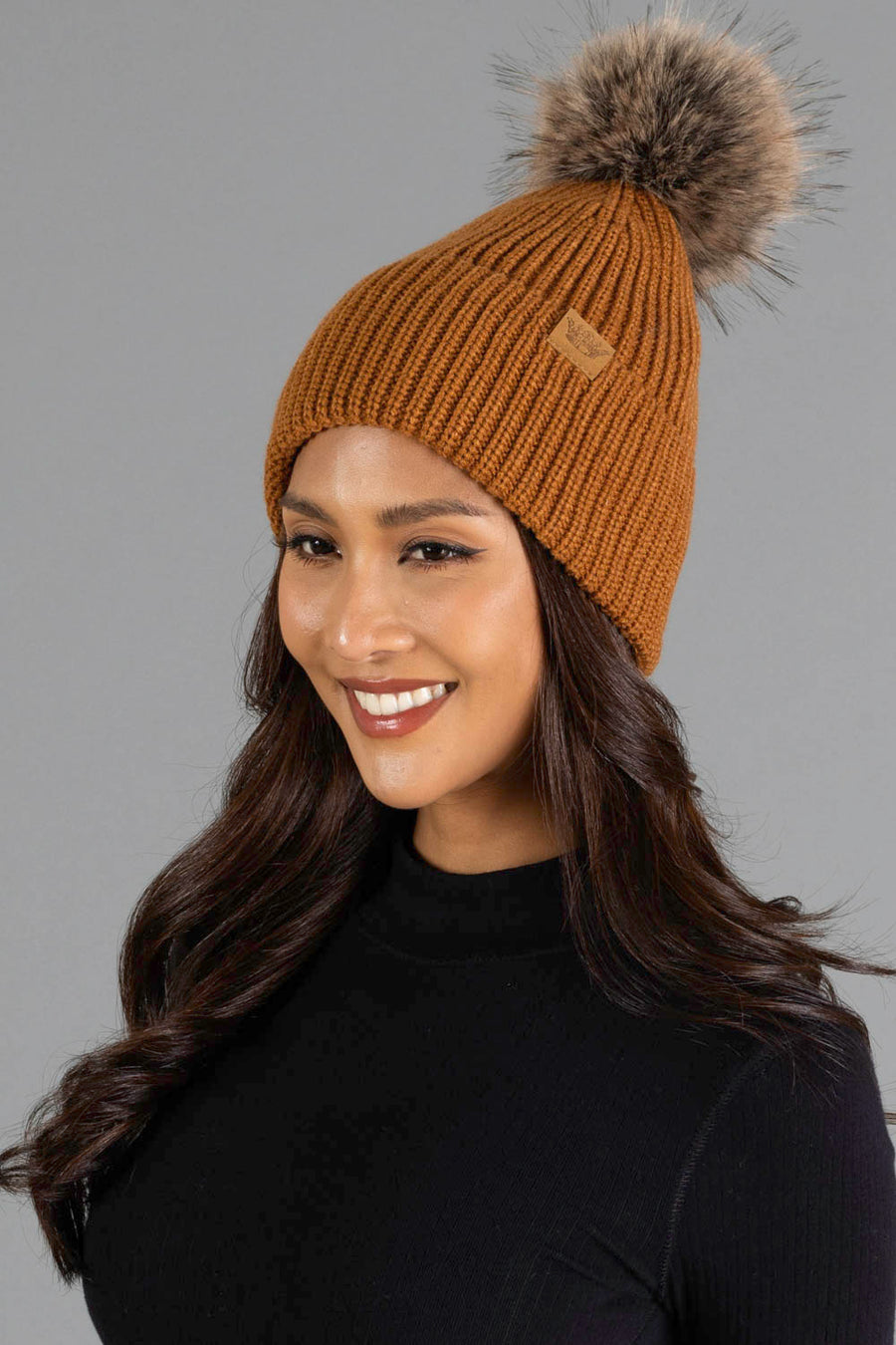 A woman with long dark hair, wearing a black top and the Camel Knit Pom Hat by Panache Apparel Collection, a brown knit beanie featuring a faux fur pom-pom, smiles against a plain gray background.