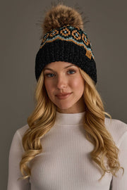 A woman with long blonde hair wears the Panache Apparel Collection’s Black & Multicolored Patterned Chenille Pom Hat, featuring a large fur pom-pom, paired with a white ribbed turtleneck sweater against a plain background.