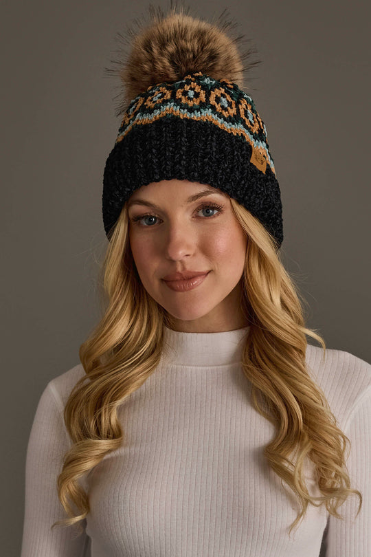 A woman with long blonde hair wears the Panache Apparel Collection’s Black & Multicolored Patterned Chenille Pom Hat, featuring a large fur pom-pom, paired with a white ribbed turtleneck sweater against a plain background.