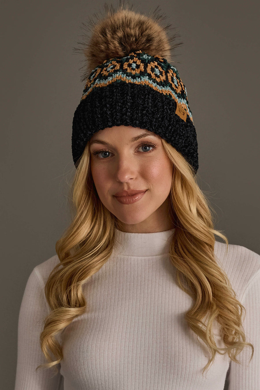A woman with long blonde hair wears the Panache Apparel Collection’s Black & Multicolored Patterned Chenille Pom Hat, featuring a large fur pom-pom, paired with a white ribbed turtleneck sweater against a plain background.
