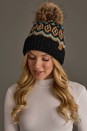A woman with long blonde hair wears the Panache Apparel Collection Black & Multicolored Patterned Chenille Pom Hat, featuring a faux fur pom-pom. She is smiling slightly and dressed in a white ribbed turtleneck against a neutral gray background.
