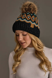 A woman with long blonde hair wears the Panache Apparel Collection Black & Multicolored Patterned Chenille Pom Hat, featuring a geometric design and a faux fur pom-pom, paired with a white ribbed turtleneck. She looks down and smiles softly against a neutral background.