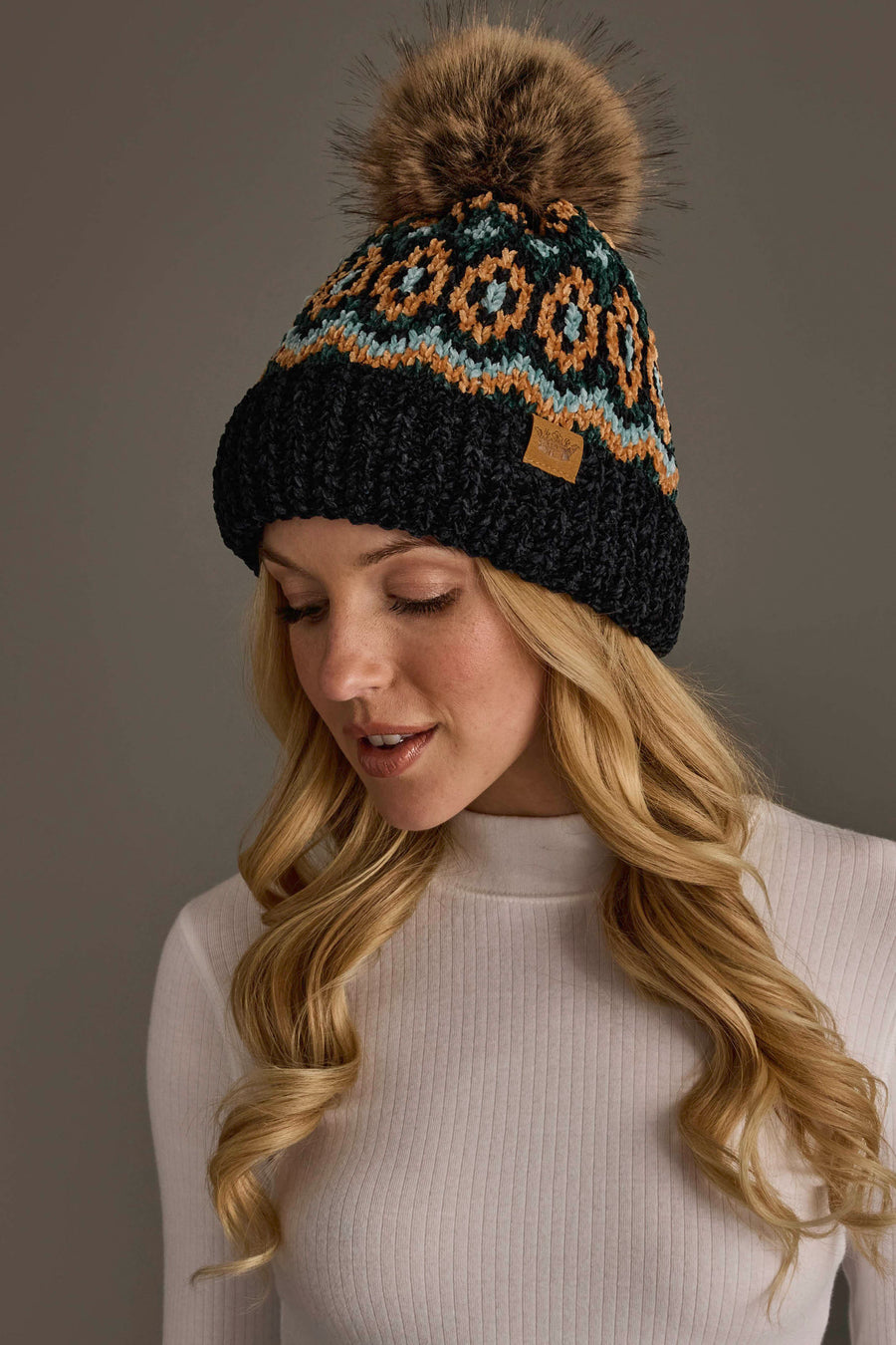 A woman with long blonde hair wears the Panache Apparel Collection Black & Multicolored Patterned Chenille Pom Hat, featuring a geometric design and a faux fur pom-pom, paired with a white ribbed turtleneck. She looks down and smiles softly against a neutral background.