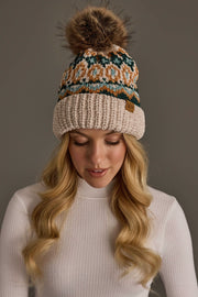 A woman with long blonde hair, wearing a white ribbed sweater and the Beige & Multicolored Patterned Chenille Pom Hat from Panache Apparel Collection, which features a large faux fur pom-pom, looks down against a plain gray background.
