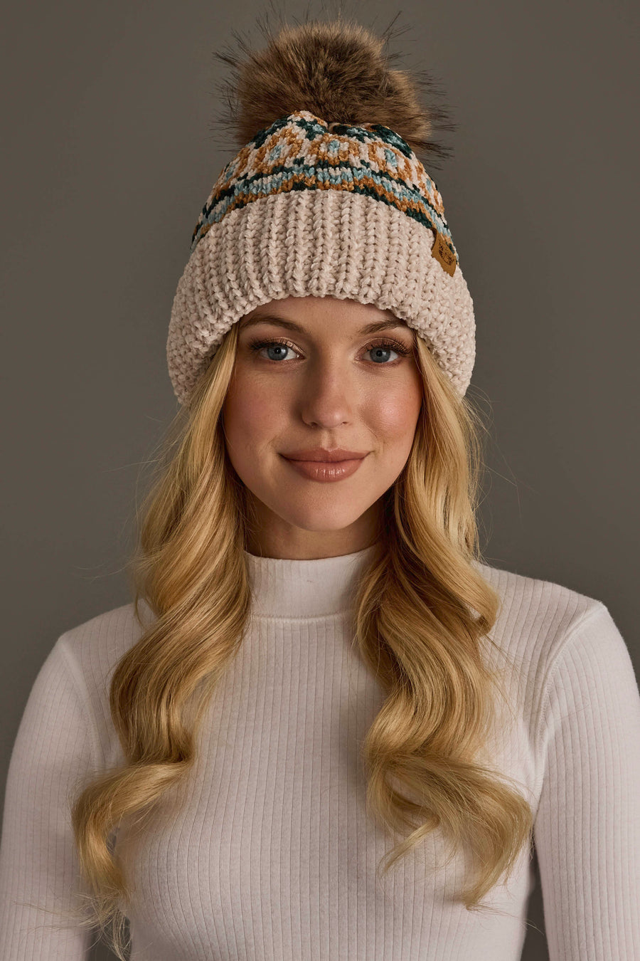 A woman with long blonde hair is smiling in front of a plain gray background, wearing a white ribbed turtleneck and the Panache Apparel Collection’s Beige & Multicolored Patterned Chenille Pom Hat.