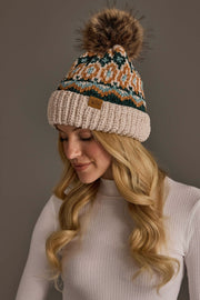 A woman with long blonde hair is wearing a white ribbed turtleneck sweater and the Beige & Multicolored Patterned Chenille Pom Hat by Panache Apparel Collection, looking down and smiling softly against a plain background.