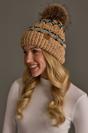 A woman with long blonde hair is smiling against a plain background while wearing the Panache Apparel Collection’s Tan & Multicolored Patterned Chenille Pom Hat and a white ribbed turtleneck sweater.