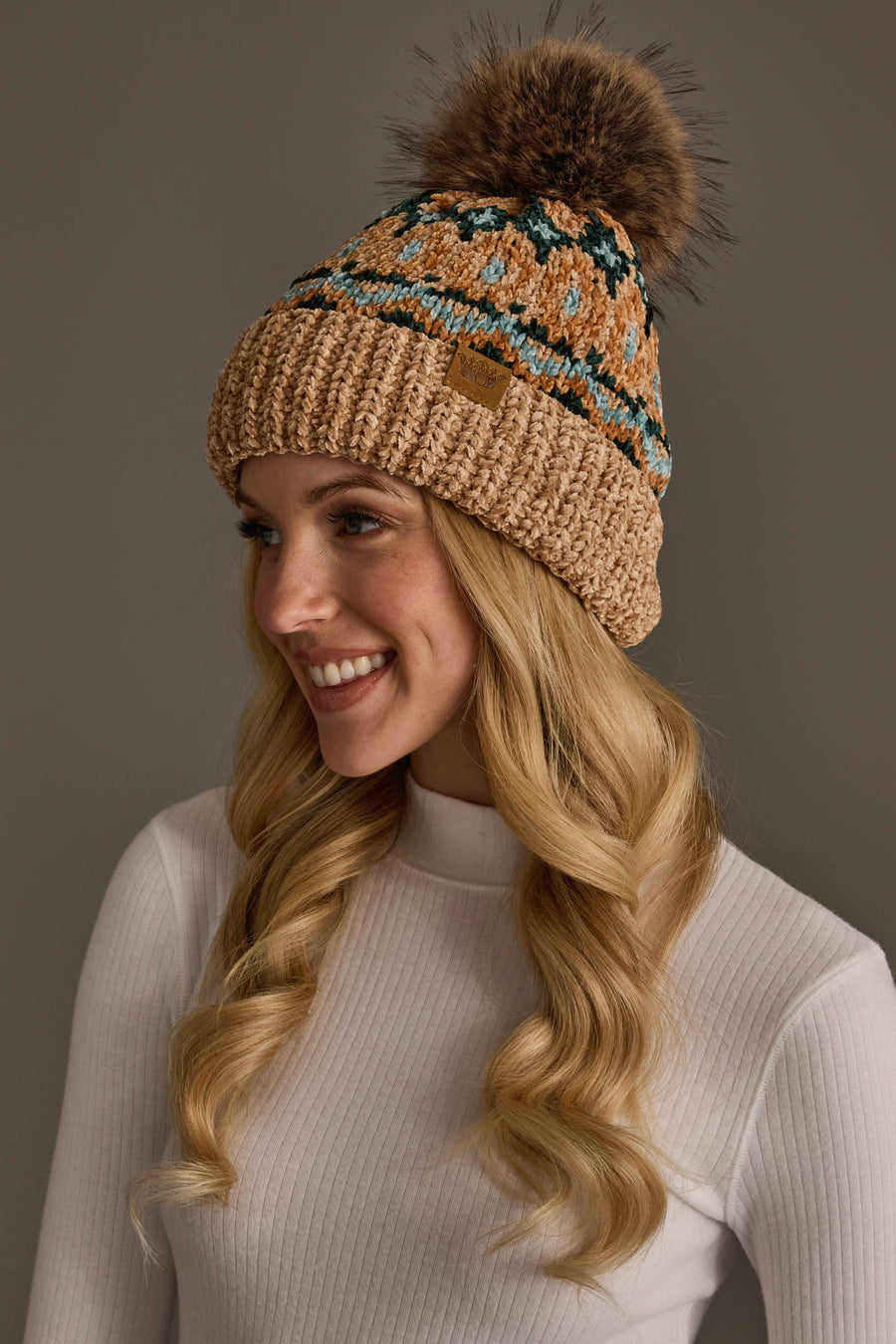 A woman with long blonde hair is smiling against a plain background while wearing the Panache Apparel Collection’s Tan & Multicolored Patterned Chenille Pom Hat and a white ribbed turtleneck sweater.
