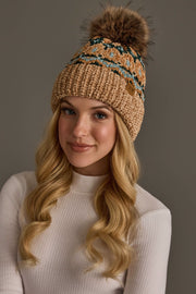 A woman with long blonde hair wears the Tan & Multicolored Patterned Chenille Pom Hat by Panache Apparel Collection, a knitted tan beanie featuring blue-green patterns and a fur pom-pom, paired with a white ribbed long-sleeve top, standing against a plain gray background.