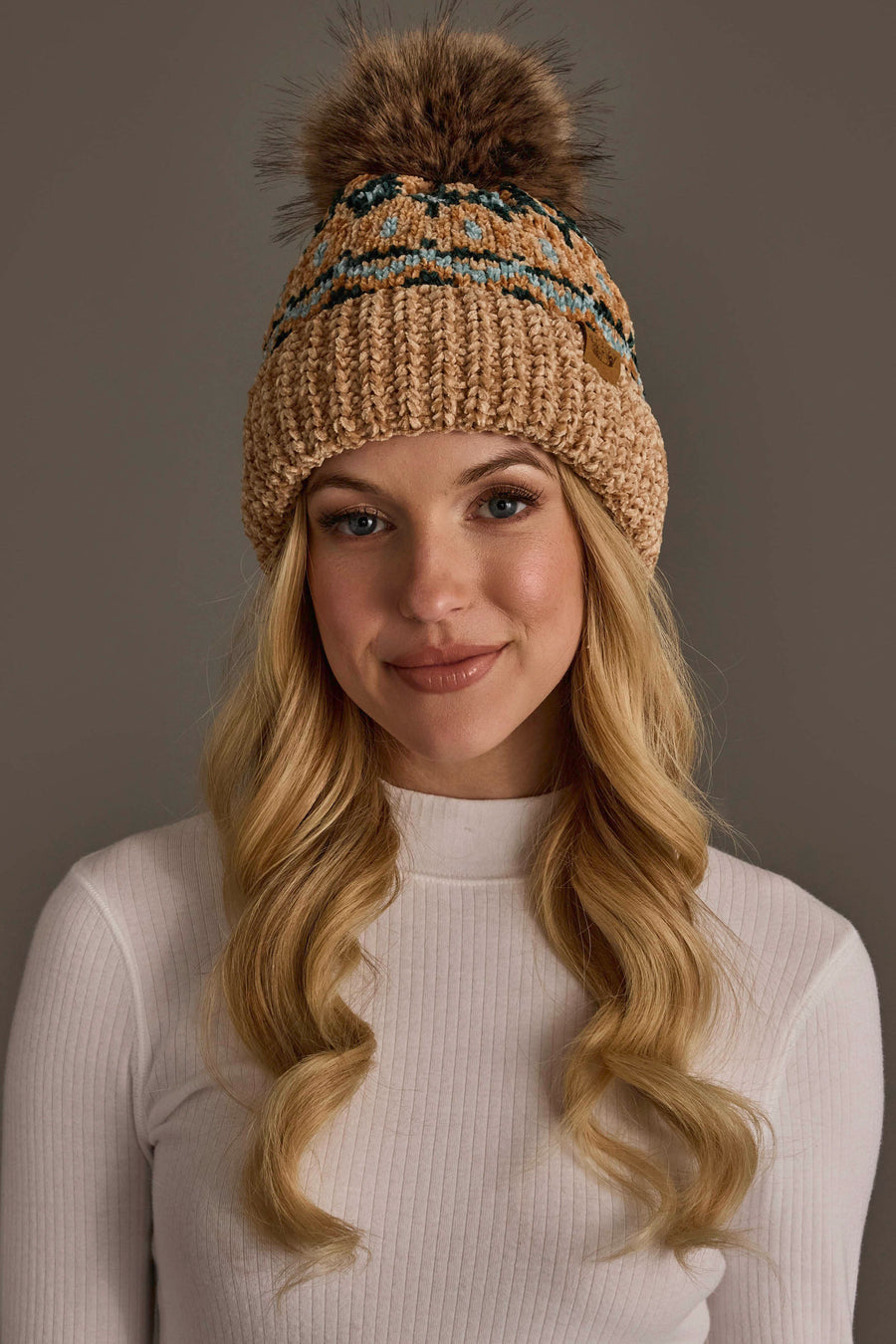 A woman with long blonde hair wears the Panache Apparel Collection’s Tan & Multicolored Patterned Chenille Pom Hat and a white ribbed shirt, standing against a neutral background.