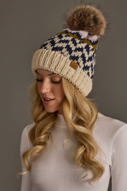 A woman with long blonde hair wears the Beige, Navy & Lilac Patterned Pom Hat from Panache Apparel Collection, paired with a white ribbed sweater. She is looking down and smiling slightly against a plain gray background.