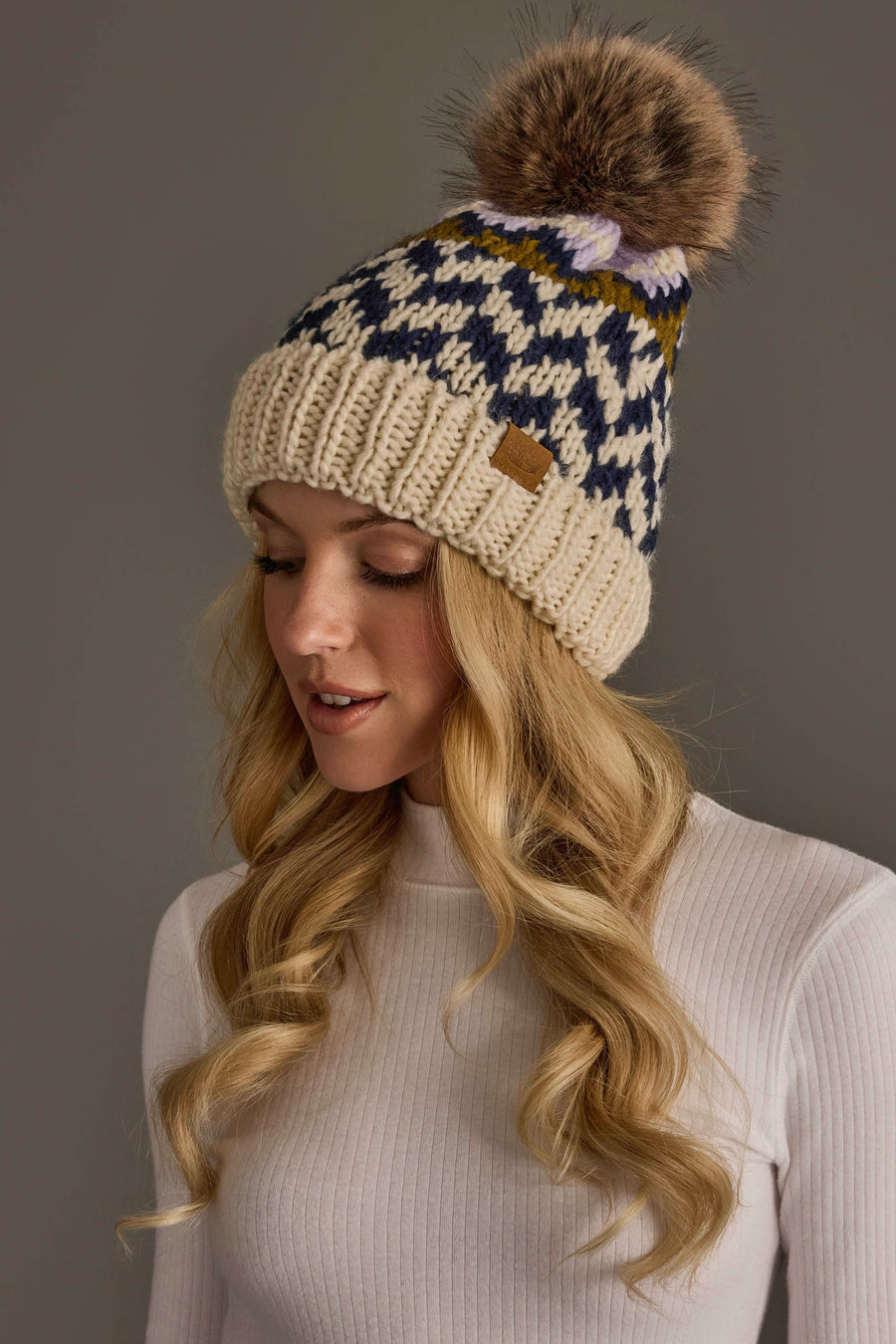 A woman with long blonde hair wears the Beige, Navy & Lilac Patterned Pom Hat from Panache Apparel Collection, paired with a white ribbed sweater. She is looking down and smiling slightly against a plain gray background.