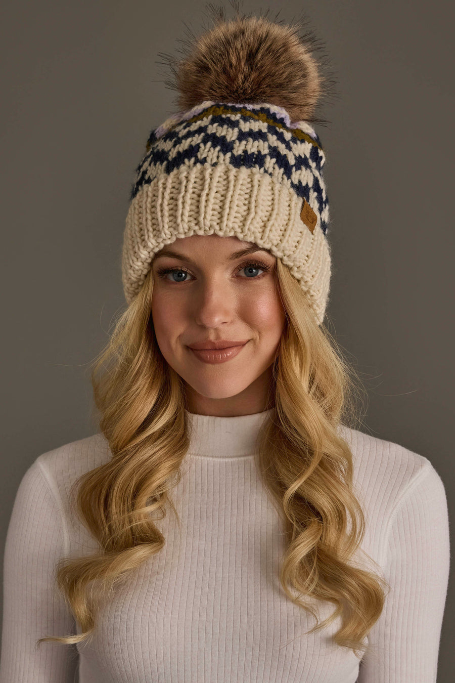 A woman with long blonde hair wears a white ribbed sweater and the Panache Apparel Collection Beige, Navy & Lilac Patterned Pom Hat, featuring a large faux fur pom-pom, as she poses against a plain gray background.
