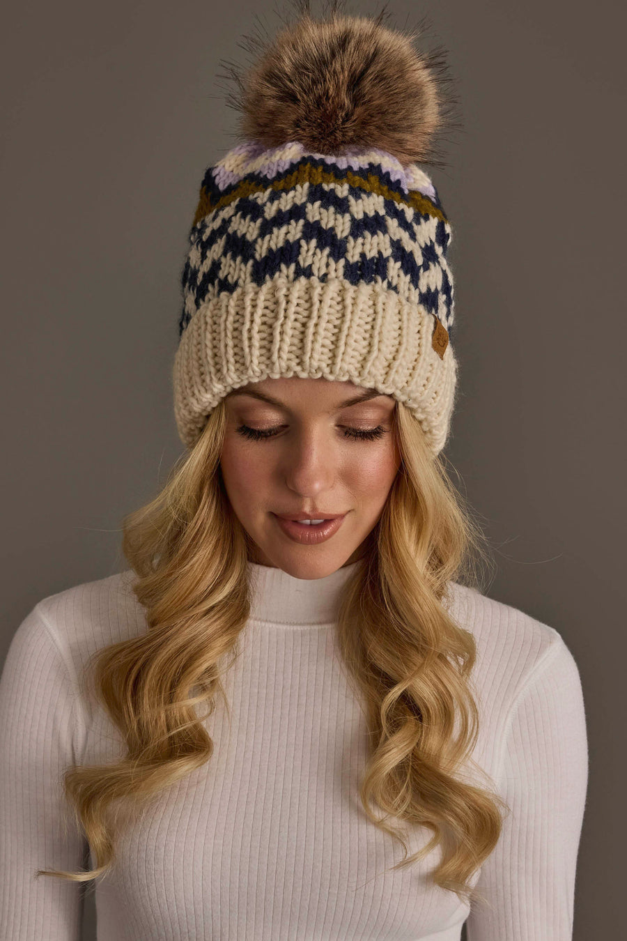 A woman with long blonde hair wears a white ribbed sweater and the Panache Apparel Collection Beige, Navy & Lilac Patterned Pom Hat, featuring a knitted geometric design and a large faux fur pom-pom on top. She is looking down with a neutral expression.