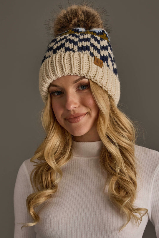 A woman with long blonde hair, smiling softly against a neutral background, wears a white ribbed shirt and the Panache Apparel Collection Beige, Navy & Light Blue Patterned Pom Hat.