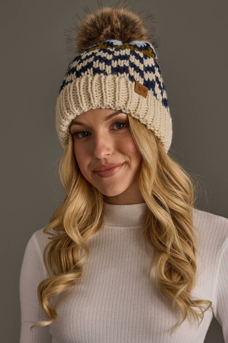 A woman with long blonde hair, smiling softly against a neutral background, wears a white ribbed shirt and the Panache Apparel Collection Beige, Navy & Light Blue Patterned Pom Hat.