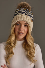 A woman with long blonde hair wears the Panache Apparel Collection Beige, Navy & Light Blue Patterned Pom Hat, featuring a faux fur pom, along with a light ribbed turtleneck shirt, standing against a plain gray background.