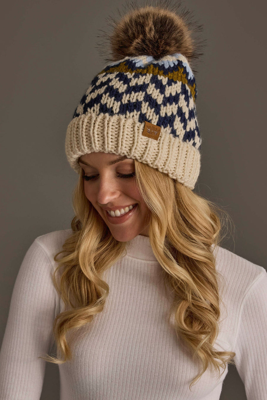 A woman with long blonde hair wears a white ribbed sweater and the Panache Apparel Collection Beige, Navy & Light Blue Patterned Pom Hat, smiling as she looks down against a plain background.
