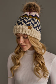 A woman with long blonde hair wears a white ribbed sweater and the Beige, Navy & Light Pink Patterned Pom Hat from Panache Apparel Collection, featuring a blue, white, pink, and mustard design with a pom-pom. She is looking down against a plain background.