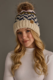 A woman with long blonde hair wears the Panache Apparel Collection’s Beige, Navy & Light Pink Patterned Pom Hat, with a fur pom-pom, paired with a white ribbed sweater, standing against a plain gray background.