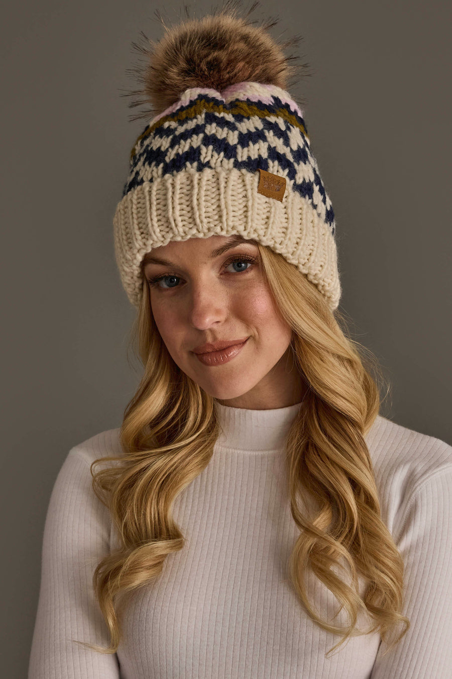 A woman with long blonde hair wears the Panache Apparel Collection’s Beige, Navy & Light Pink Patterned Pom Hat, with a fur pom-pom, paired with a white ribbed sweater, standing against a plain gray background.