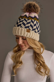 A woman in a white ribbed sweater wears the Panache Apparel Collection’s Beige, Navy & Light Pink Patterned Pom Hat, featuring a large brown pom-pom, as she looks down and smiles.