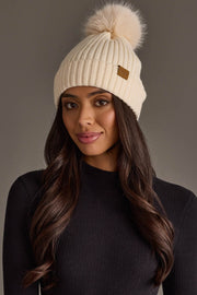 A woman with long brown hair wears the Panache Apparel Collection Cream Knit Hat with Cream Pom along with a black ribbed sweater, standing against a neutral background.