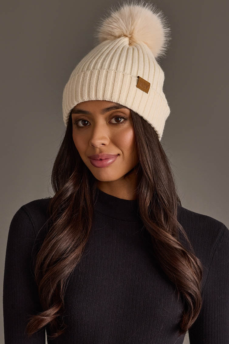 A woman with long brown hair wears the Panache Apparel Collection Cream Knit Hat with Cream Pom along with a black ribbed sweater, standing against a neutral background.
