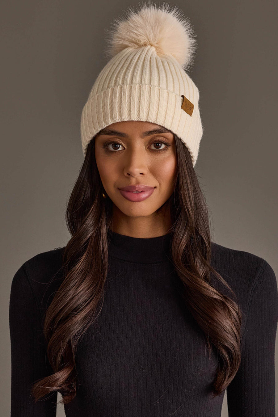 A woman with long brown hair is wearing the Panache Apparel Collection Cream Knit Hat with Cream Pom and a black ribbed sweater, standing against a plain gray background.