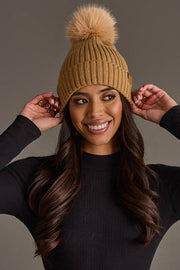 A smiling woman wearing the Panache Apparel Collection Tan Knit Hat with Tan Pom, styled with a black long-sleeve top and her long wavy hair, poses against a plain background with her hands touching the sides of her hat.