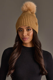 A woman is wearing the Panache Apparel Collection Tan Knit Hat with Tan Pom and a black ribbed long-sleeve shirt, standing against a plain gray background.
