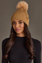 A woman wearing the Panache Apparel Collection Tan Knit Hat with Tan Pom, paired with a black ribbed sweater and her long wavy brown hair, stands against a neutral background.