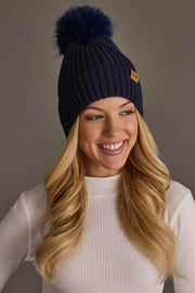 A smiling woman with long blonde hair wears the Panache Apparel Collection’s Dark Navy Knit Hat with Navy Pom, featuring a large pom-pom and small brown tag, paired with a white ribbed sweater against a plain gray background.