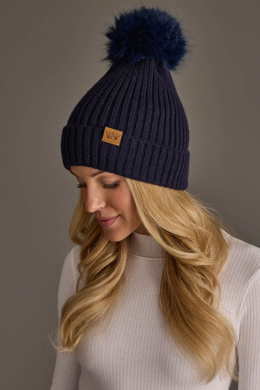 A woman with long blonde hair wears the Dark Navy Knit Hat with Navy Pom by Panache Apparel Collection. She looks down with a slight smile, dressed in a light ribbed sweater against a neutral background.
