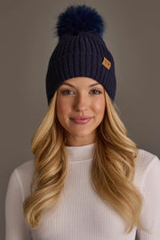 A woman with long blonde hair, wearing a white ribbed turtleneck and the Panache Apparel Collection Dark Navy Knit Hat with Navy Pom, smiles gently against a plain background.
