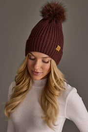A woman with long blonde hair is wearing the Panache Apparel Collection Brown Knit Hat with Brown Pom and a white ribbed turtleneck sweater against a neutral background.