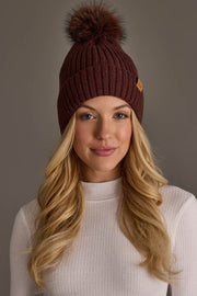 A woman with long blonde hair wears a white ribbed turtleneck sweater and the Brown Knit Hat with Brown Pom from Panache Apparel Collection. She is facing the camera and smiling slightly against a plain gray background.
