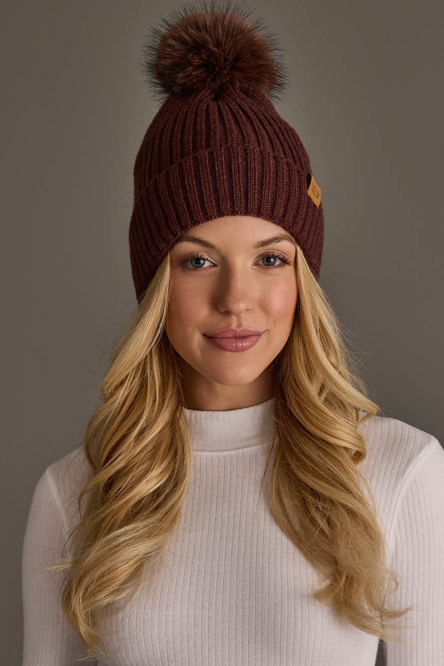 A woman with long blonde hair wears a white ribbed turtleneck sweater and the Brown Knit Hat with Brown Pom from Panache Apparel Collection. She is facing the camera and smiling slightly against a plain gray background.