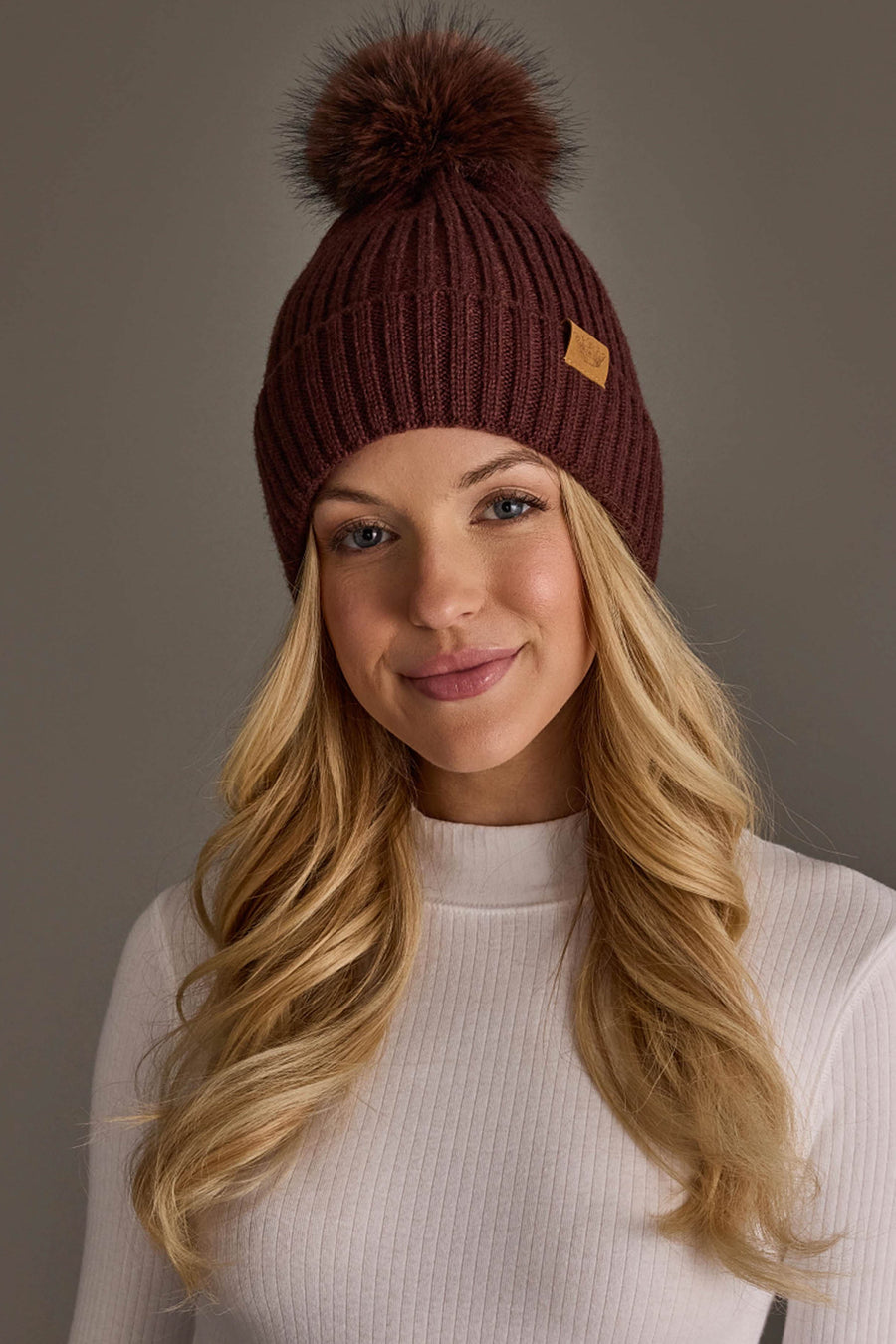 A woman with long blonde hair is wearing the Panache Apparel Collection Brown Knit Hat with Brown Pom and a white ribbed sweater, standing against a plain gray background.