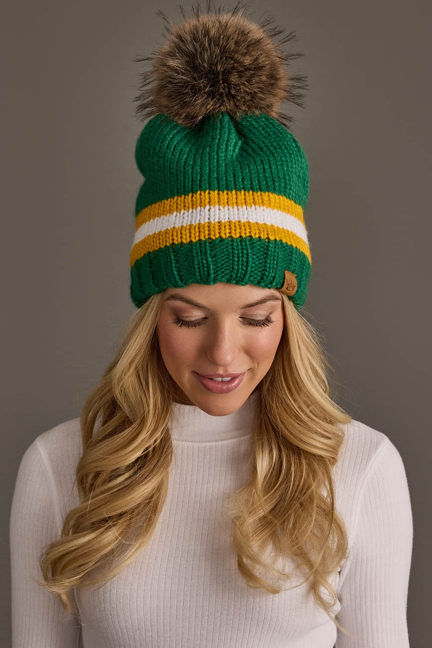 A woman wearing the Panache Apparel Collection Green, Gold & White Knit Pom Hat, featuring yellow and white stripes and a brown pom-pom, looks down while dressed in a white ribbed turtleneck sweater against a plain background.