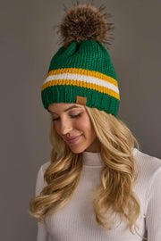 A woman smiles while looking down against a plain gray background, wearing the Panache Apparel Collection’s Green, Gold & White Knit Pom Hat featuring yellow and white stripes with a brown pom-pom paired with a white ribbed turtleneck.